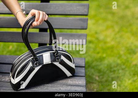 Sac à main noir sur le banc de stationnement. Femme tenant un sac à main noir et blanc sur un banc de parc en bois avec fond d'herbe verte et ambiance de style de vie en plein air. Banque D'Images