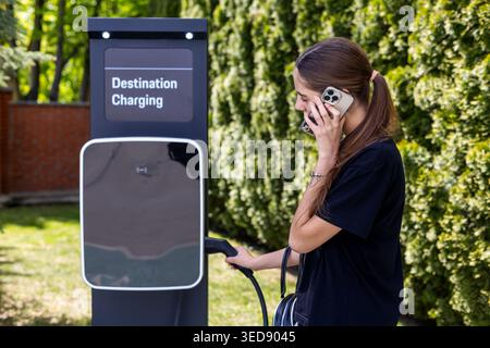 Femme appelant tout en utilisant EV charger. Jeune femme parle sur un smartphone tout en tenant un câble de recharge de véhicule électrique à une destination extérieure Banque D'Images