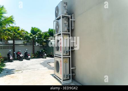 Cette image montre plusieurs unités de climatisation extérieures installées le long d'un mur, avec de la verdure environnante et un parking avec des motos, illu Banque D'Images
