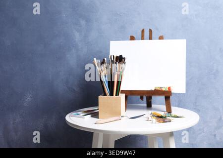 Pinceaux, couteau, peintures et chevalet avec toile sur table près du mur bleu. Espace pour le texte Banque D'Images