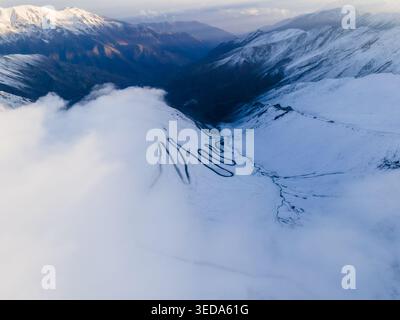 Vue aérienne de la route sinueuse de Babusar Pass à travers des sommets de montagne enneigés et des nuages blancs épais à Babusar, KPK, Pakistan. Banque D'Images
