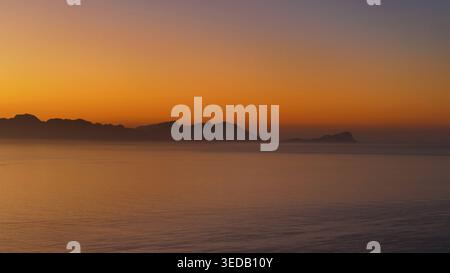 Vue aérienne de la chaîne de montagnes silhouettée à travers les eaux calmes de l'océan sous un ciel de coucher de soleil orange et violet vibrant à Cape Town, Cap occidental, SO Banque D'Images