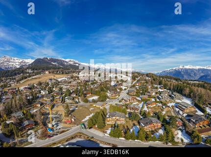 Vue aérienne de la station balnéaire de Crans-Montana avec chalets traditionnels, arbres à feuilles persistantes et sommets alpins enneigés sous un ciel bleu clair de Crans-Montana, Banque D'Images