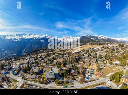 Vue aérienne du village alpin avec chalets traditionnels et pins sur fond de sommets enneigés et d'un ciel bleu clair à Crans-Mont Banque D'Images