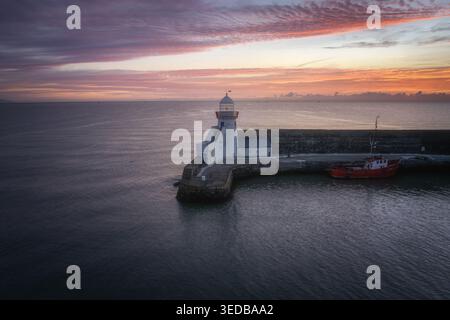 Vue aérienne du phare de Balbriggan au bout d'une jetée de pierre avec un bateau rouge amarré sous un ciel de coucher de soleil vibrant Balbriggan, comté de Dublin, Irlande. Banque D'Images