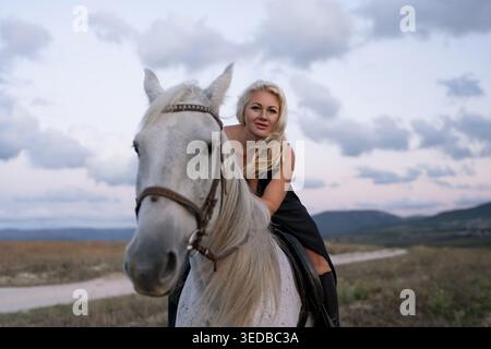 Femme cheval blonde cavalier élégant assis sur un cheval blanc majestueux au coucher du soleil Banque D'Images