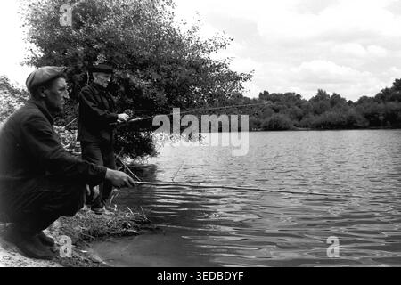 Région de Donetsk, années 1960, sur la rive. Deux hommes locaux sont engagés dans la pêche amateur de la côte sablonneuse. Le pêcheur au premier plan accroupit bas, tenant une longue tige de bambou au-dessus de l'eau, tandis que son compagnon se tient derrière lui avec une tige de bobine. Entourés d'un dense feuillage estival, ils profitent d'un après-midi tranquille de loisirs en plein air. Ce cliché noir et blanc franc présente les passe-temps ruraux soviétiques traditionnels et les loisirs masculins. Une véritable archive historique reflétant la vie calme et paisible d'avant la guerre. Banque D'Images