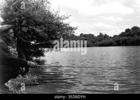 Région de Donetsk, années 1960, sur la rive. Deux hommes locaux sont engagés dans la pêche amateur de la côte sablonneuse. Le pêcheur au premier plan accroupit bas, tenant une longue tige de bambou au-dessus de l'eau, tandis que son compagnon se tient derrière lui avec une tige de bobine. Entourés d'un dense feuillage estival, ils profitent d'un après-midi tranquille de loisirs en plein air. Ce cliché noir et blanc franc présente les passe-temps ruraux soviétiques traditionnels et les loisirs masculins. Une véritable archive historique reflétant la vie calme et paisible d'avant la guerre. Banque D'Images