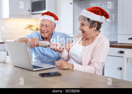 Ordinateur portable, vieux couple et cracker festif pour Noël, bonheur et appel vidéo pour la célébration. Amour, décoration ou homme senior en cuisine, femme ou Banque D'Images