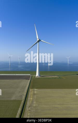 Vue aérienne d'une grande éolienne blanche debout sur des champs agricoles verts avec un parc éolien offshore dans la mer bleue sous un ciel dégagé. Banque D'Images