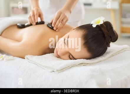 Femme cliente couchée paisiblement sur la table de massage blanche, recevant un massage aux pierres chaudes, mains thérapeutes Banque D'Images
