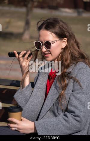 Jeune femme portant des lunettes de soleil et un manteau gris assis sur un banc dans un parc, tenant une tasse à café et enregistrant un message vocal sur son smartphone, enj Banque D'Images