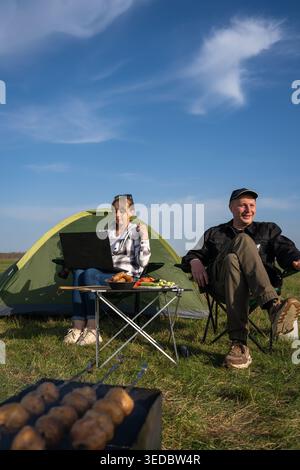 Couple camping dans une clairière verte : femme travaillant sur un ordinateur portable à une table de pique-nique tandis que l'homme se détend par un barbecue, profitant du plein air ensoleillé, travail à distance et Banque D'Images