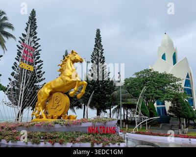 Une grande statue de cheval d'or se tient sur un piédestal couvert de fleurs à Nha Trang Vietnam derrière elle est la tour Tram Huong en forme de lotus blanc et la grande broche Banque D'Images