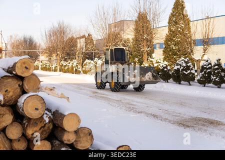 Chargeur frontal déneigeant une route devant un bâtiment d'usine, avec des bûches de bois recouvertes de neige empilées à proximité Banque D'Images
