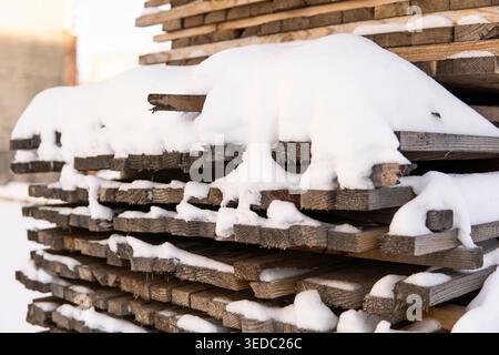 Planches de bois empilées recouvertes de neige fraîche montrant des matériaux de travail du bois pour la construction en hiver Banque D'Images