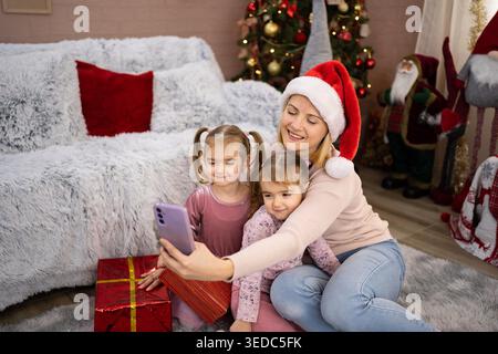 Mère portant chapeau de Père Noël prenant selfie avec ses deux filles près de l'arbre de Noël et des cadeaux Banque D'Images