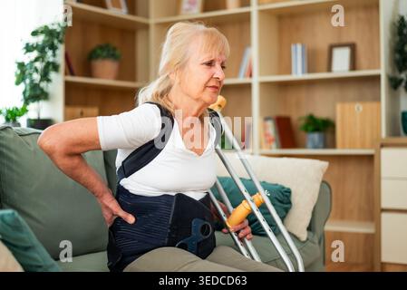 La femme âgée portant une attelle de soutien du dos est assise sur le lit à la maison. Elle a mal au dos. Banque D'Images