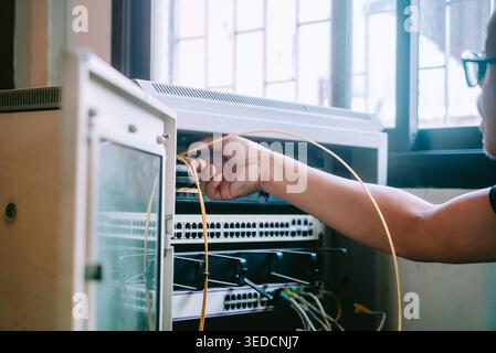 Un technicien effectue la configuration de l'installation réseau dans un bureau. L'image capture une main connectant des câbles dans une armoire d'équipement réseau, showcas Banque D'Images