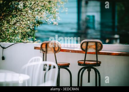 Un cadre extérieur serein avec deux tabourets de bar confortables sur un patio, mis en valeur par la lumière chaude du soleil et un fond vert luxuriant, idéal pour la détente. Banque D'Images