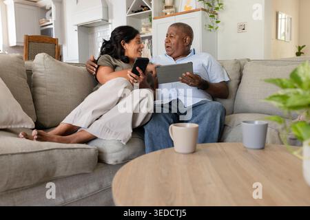 Couple senior assis près sur le canapé sectionnel gris clair à la maison tenant smartphone et tablette Banque D'Images