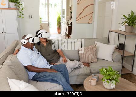 Couple aîné afro-américain assis sur le canapé portant des casques VR blancs avec des tasses à café à la maison Banque D'Images