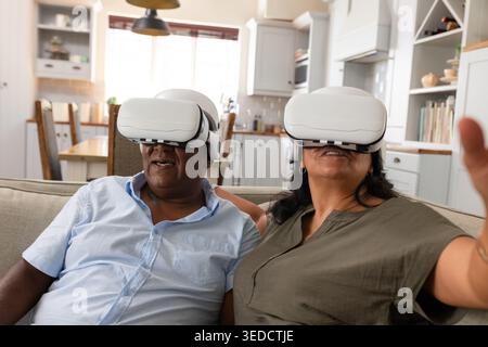 Couple senior afro-américain portant des casques VR blancs assis sur le canapé à la maison atteignant vers l'extérieur Banque D'Images