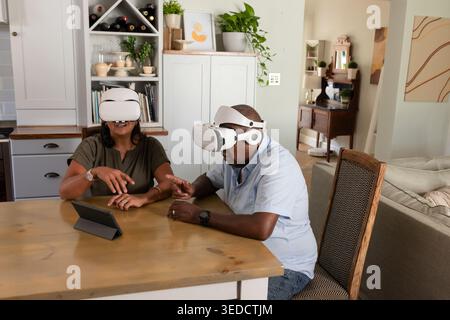 Couple afro-américain portant des casques VR blancs et interagissant avec une tablette à la table à manger à la maison Banque D'Images