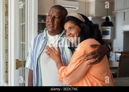 Couple aîné afro-américain debout embrassant dans la cuisine par la porte vitrée, montrant des bagues et la montre Banque D'Images