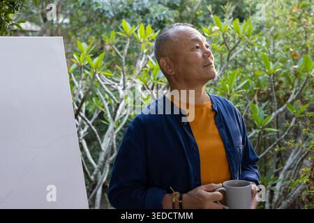 Homme asiatique senior debout dans le jardin tenant une tasse grise portant un tee-shirt orange bleu foncé chemise par toile Banque D'Images