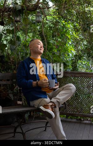 Homme asiatique senior assis sur un banc en bois sur un porche couvert tenant une tasse en céramique, portant une chemise marine Banque D'Images