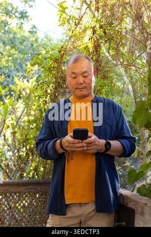 Homme asiatique senior portant un tee-shirt orange et une chemise bleue debout sur le porche vérifiant smartphone par treillis Banque D'Images