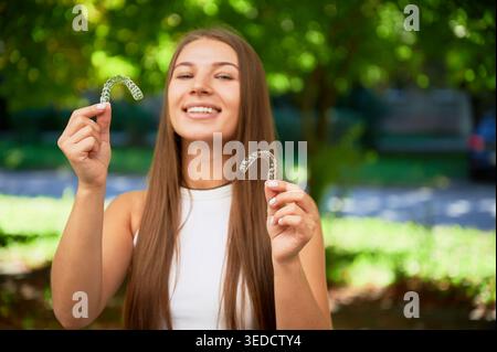 La jeune femme montre des aligneurs dentaires clairs et sourit à l'extérieur. Portrait naturel avec dents saines et plateaux orthodontiques. Idéal pour la publicité orthodontique, le contenu des soins buccaux. Focus sur les aligneurs dentaires Banque D'Images