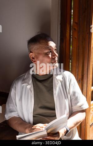 Homme asiatique senior assis près de la fenêtre en bois, portant une chemise olive, tenant un livre ouvert, regarder visible Banque D'Images