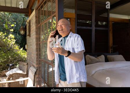 Homme asiatique senior debout dans la porte de la chambre à coucher au patio, souriant, parlant au téléphone, tenant du verre Banque D'Images
