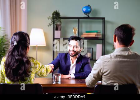 Couple indien discutant avec l'agent à la maison pendant la réunion d'investissement avec l'homme d'affaires examinant les documents financiers et les dossiers tout en étant assis sur le canapé dedans Banque D'Images
