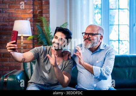Jeune fils indien et père aîné utilisant un smartphone pour les appels vidéo tout en étant assis sur le canapé dans la maison moderne, souriant et agitant pendant la conversation en ligne Banque D'Images