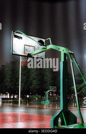 Terrain de basket nocturne avec cerceau vert et surface rouge humide, éclairé sous des lumières artificielles Banque D'Images