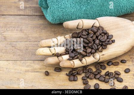 Gros plan d'un mannequin en bois qui bosse à la main des grains de café torréfiés foncés, utilisés pour exfolier la peau dans les soins de spa biologiques, avec des grains en vrac dispersés sur Banque D'Images