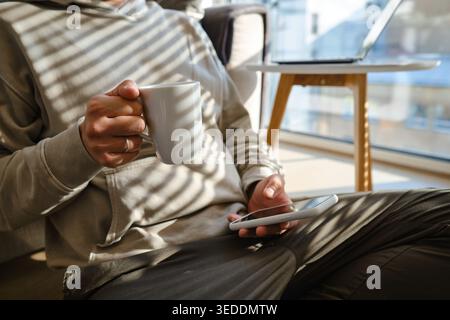Mains d'homme dans un sweat à capuche beige tenant et utilisant un smartphone avec un étui blanc près d'une fenêtre Banque D'Images