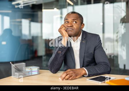 Professionnel masculin mature assis à un bureau, posant son menton sur sa main tout en regardant vers le haut, exprimant des sentiments d'ennui, de frustration et un manque de motivation pendant une longue journée de travail Banque D'Images