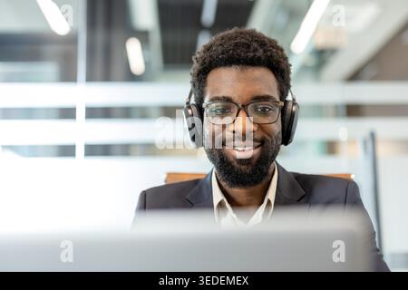 Homme professionnel souriant en toute confiance à son bureau, portant des écouteurs et des lunettes, participant à une réunion virtuelle ou à un apprentissage en ligne dans un espace de travail d'entreprise moderne Banque D'Images