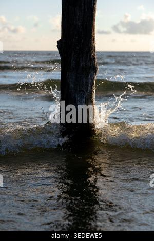 Les vagues ont frappé un poteau en bois debout dans l'eau peu profonde à la plage. Le soleil se couche en arrière-plan, projetant de la lumière sur la scène. De l'eau éclabousse autour Banque D'Images