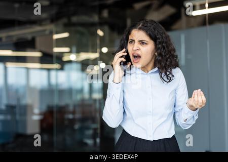 Une femme debout dans un bureau, criant dans un smartphone avec une expression angoissante et gestuelle en détresse montre la frustration, le stress, les conflits et la mauvaise communication sur le lieu de travail pendant un appel professionnel tendu Banque D'Images