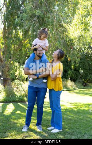 Famille afro-américaine jouant sous l'arbre tombant sur la pelouse herbeuse, homme portant l'enfant sur les épaules Banque D'Images