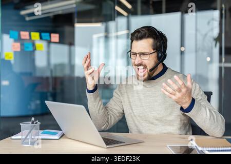 Homme frustré portant un casque et des lunettes, stressé et malheureux, criant et faisant des gestes follement sur un ordinateur portable tout en éprouvant l'épuisement professionnel dans un environnement de bureau Banque D'Images