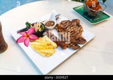 Une assiette magnifiquement agencée avec steak grillé, saucisse, frites froissées et une salade fraîche, garnie d'une fleur. Idéal pour les gourmands et resta Banque D'Images