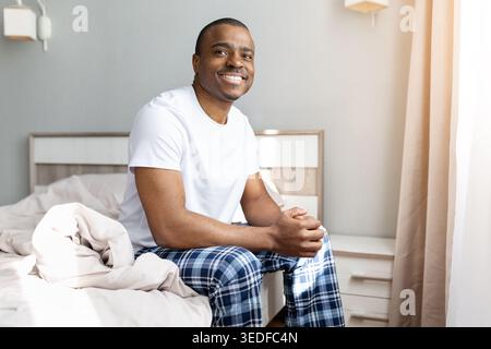 Homme afro-américain assis sur le lit souriant à la caméra dans une chambre confortable Banque D'Images