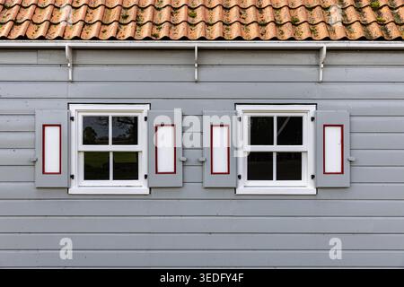 Vue sur la façade traditionnelle en bois d'un bâtiment historique avec revêtement gris et fenêtres à cadre blanc avec volets décoratifs à Schokland, FLE Banque D'Images