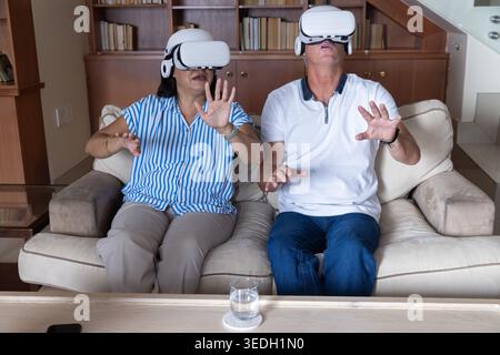 Couple senior assis sur le canapé beige portant des ensembles VR blancs, atteignant la table basse avec téléphone Banque D'Images